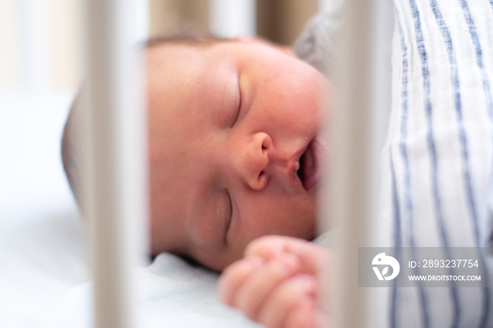 Caucasian newborn baby falling asleep in the baby bed, under striped blanket behind white slats. SID