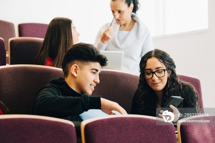 Teenage girl showing mobile phone to friend in classroom with professor in background