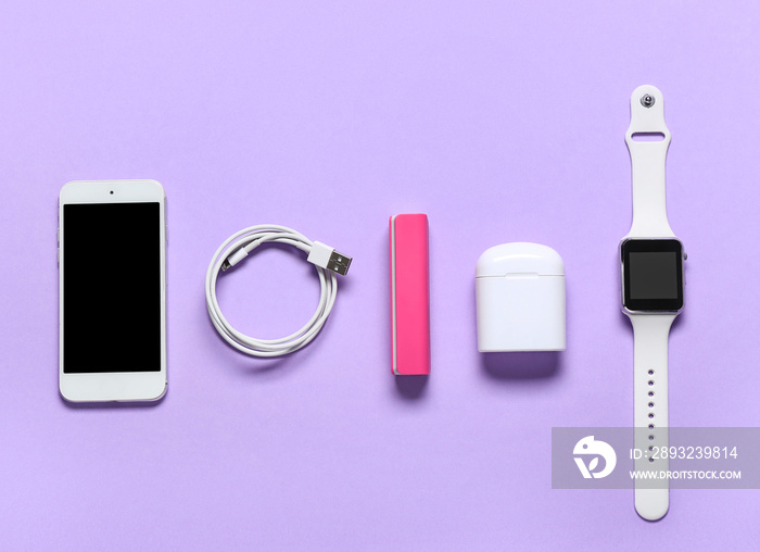 Power bank with mobile phone, USB cable, earphones and smartwatch on lilac background