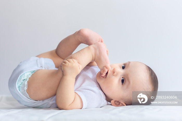 Little hungry baby boy taking feet in mouth. 6-month baby having fun in white bedding. Cute baby lyi