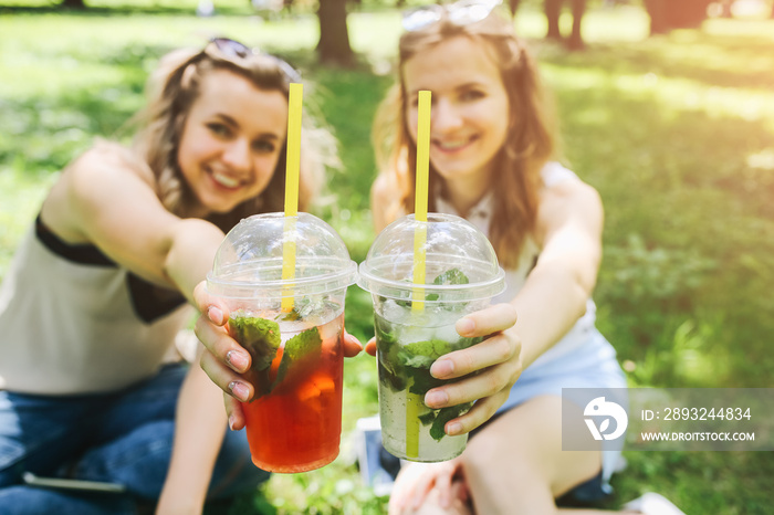 Two hipster girls are laughing and drinking summer cocktails outdoors in the green grass. Cold non-a