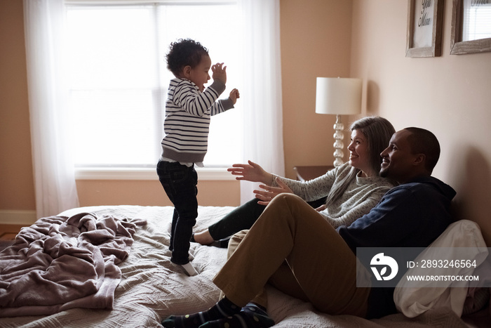 Side view of happy parents looking at cute son dancing against window on bed