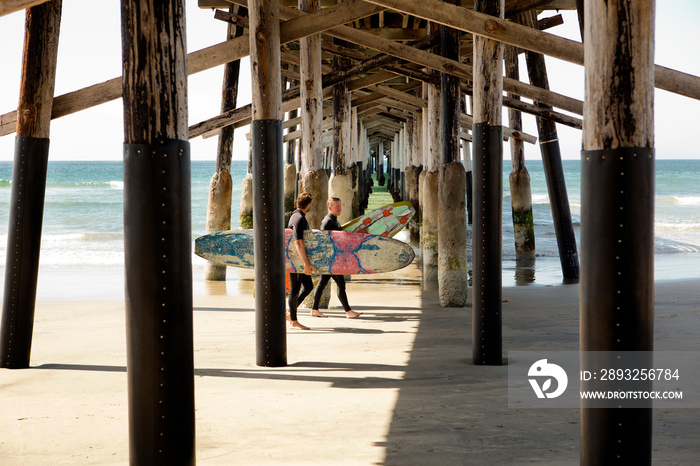 Two men walking with surfboards under pier