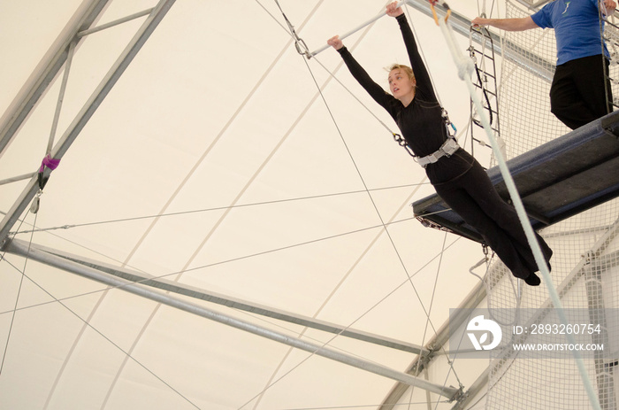 An adult female hangs on a flying trapeze at an indoor gym. The woman is an amateur trapeze artist.