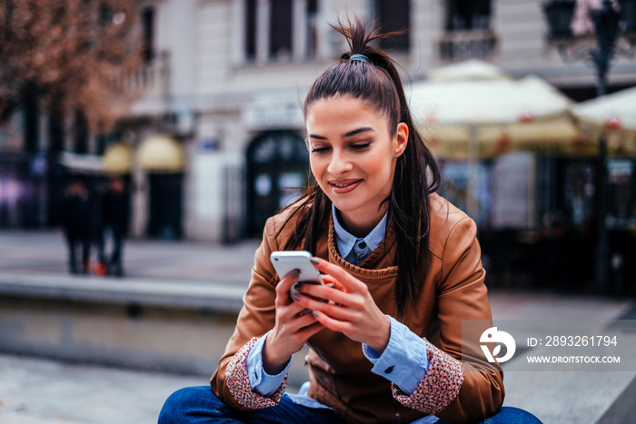 Young woman texting on smartphone while sitting at the city square.