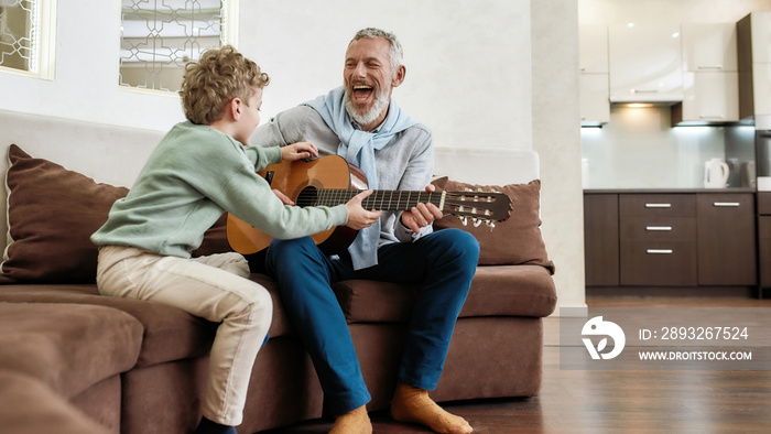 Weekend with grandpa. Cheerful mature man, grandfather playing guitar to his cute little grandson