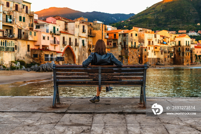 Young woman sitting on the bench and enjoying view of coastal village Cefalu at sunrise, Sicily, Ita