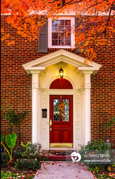 砖砌房屋的入口，门廊有柱子，人行道上有秋天的树叶和彩色的红色门