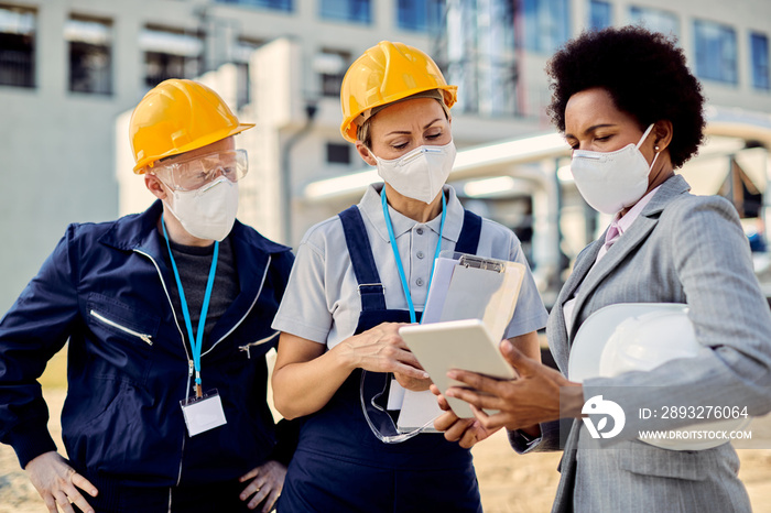 Black engineer and two workers with face masks cooperating while using digital tablet at constructio