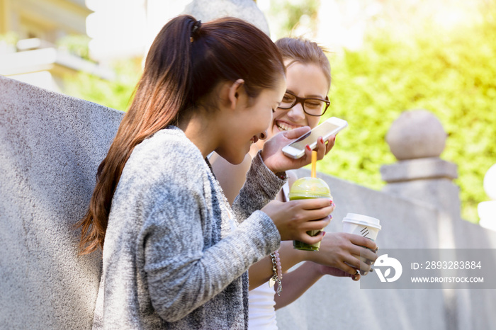 Two young women leaning against park wall laughing and drinking takeaway coffee