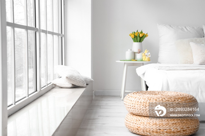 Vase with tulips, cups and Easter rabbits on table near light wall in bedroom interior