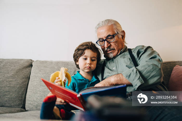 Senior man reading storybook to grandson while sitting on sofa at home