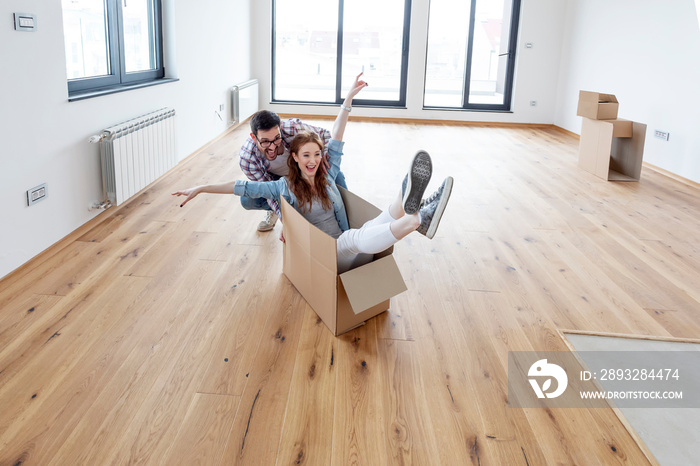 Young couple in new empty room. She is sitting on card box while he pushing her from behind