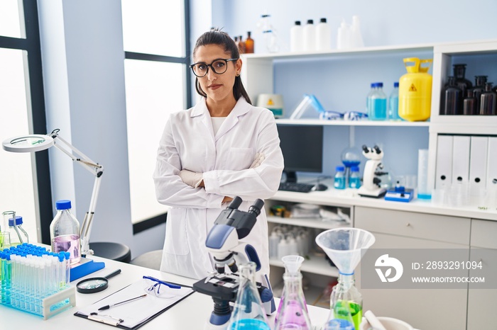 Young hispanic woman wearing scientist uniform standing with arms crossed gesture at laboratory