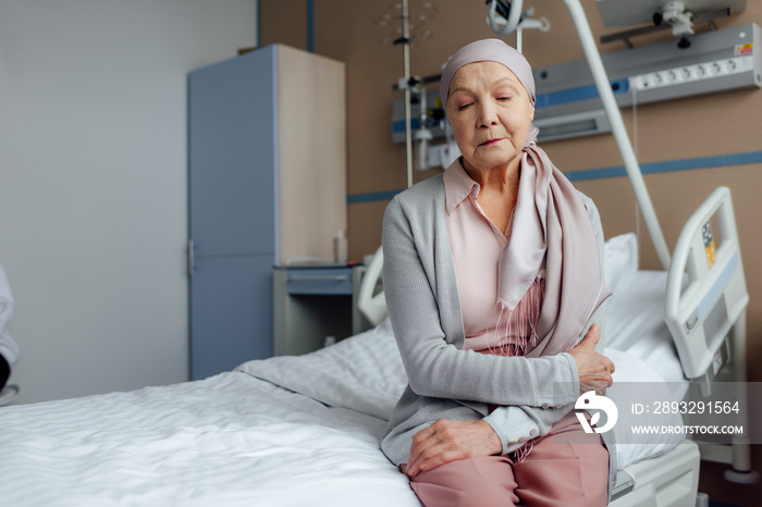 senior woman with cancer sitting on bed in hospital with hands crossed