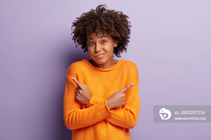 Hesitant confused black woman with Afro hairstyle, dressed in orange jumper, points at both sides, h