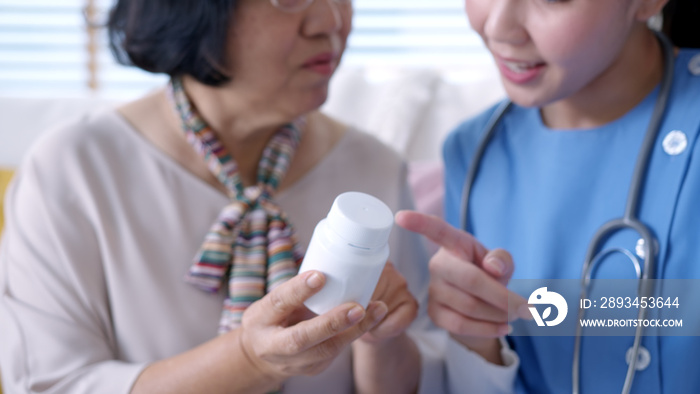 Young caregiver in scrubs uniform showing medicine bottle to old elderly asian woman in home visit c