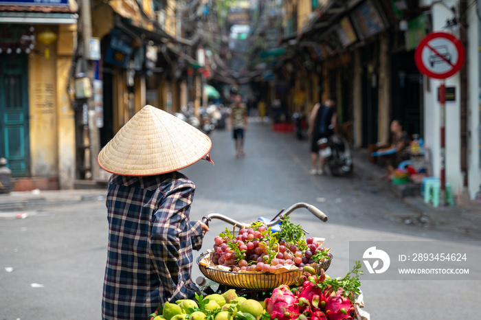 The street vendor with bike loaded of tropical fruits in old town street in Hanoi, old houses and st