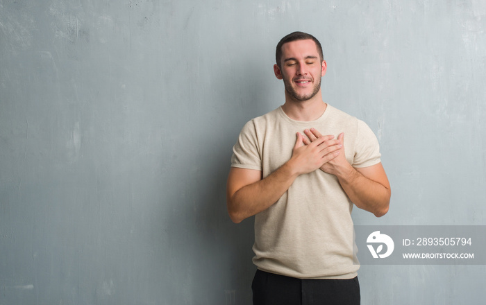 Young caucasian man over grey grunge wall smiling with hands on chest with closed eyes and grateful 