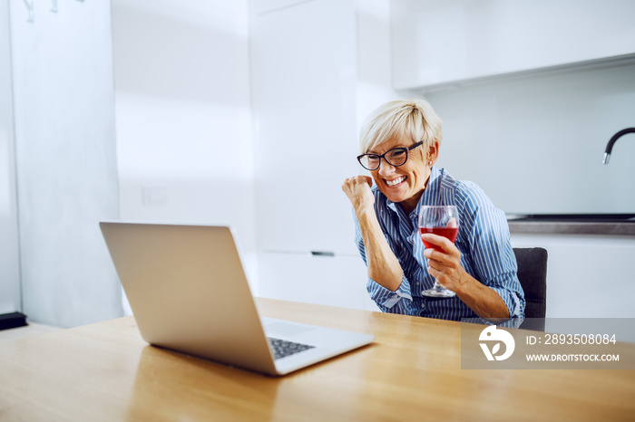 Attractive caucasian blond senior woman sitting at dining table, drinking wine, looking at laptop an