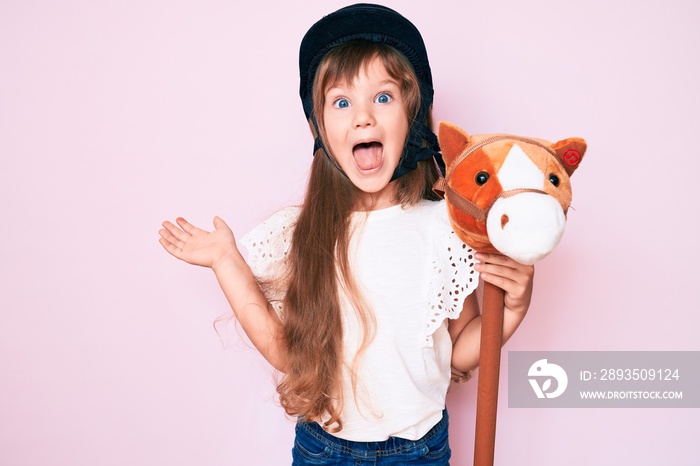 Little caucasian kid girl with long hair riding horse toy wearing vintage helmet celebrating achieve