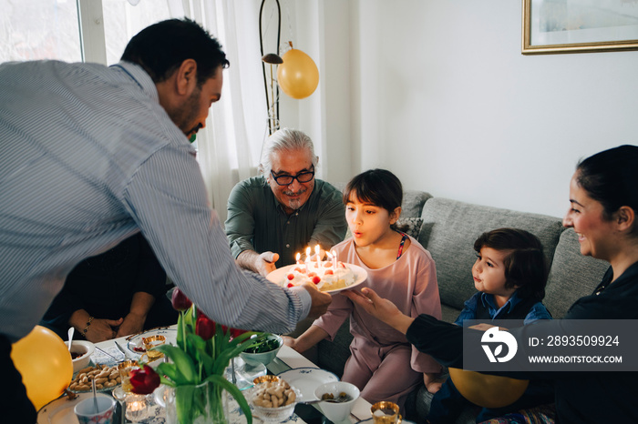 Girl blowing candles on birthday cake while sitting with family during party