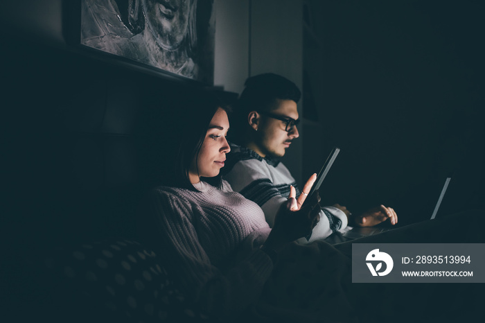 Young couple indoors at home bedtime using smartphone and tablet