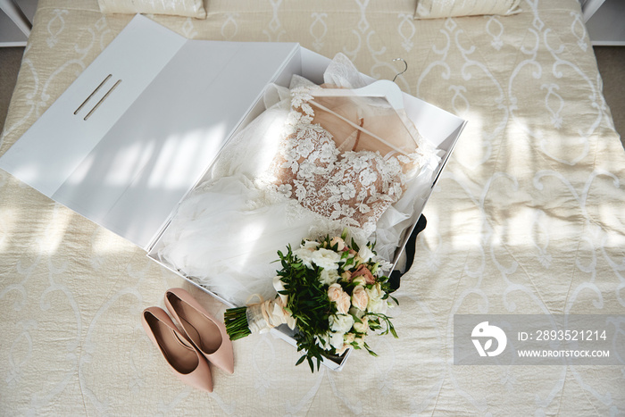 Luxury wedding dress in white box, beige womens shoes and bridal bouquet on bed, copy space. Bridal