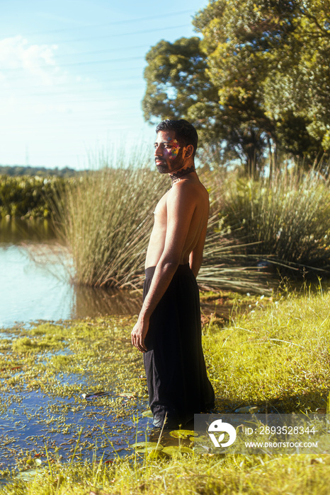 portrait of a dark skinned Indian man by a lake, shirtless, with make up, surrounded by nature