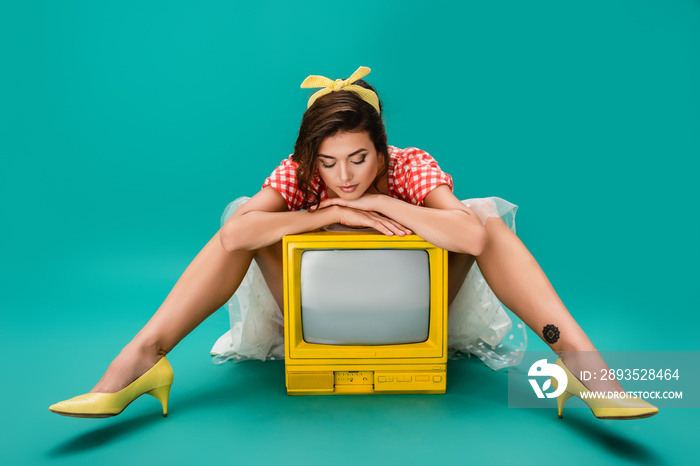 stylish pin up woman leaning on yellow retro tv set while sitting on turquoise background