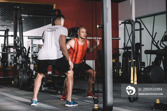 back view of personal trainer supervising young african american sportsman lifting barbell