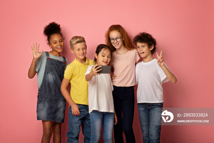 Cute diverse schoolchildren taking selfie with cellphone on pink background