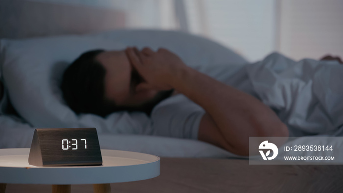 Clock on bedside table near blurred man covering face on bed at night.