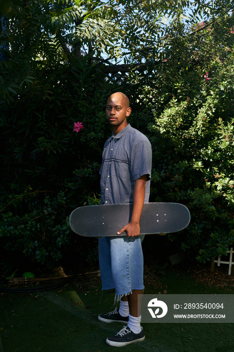 Portrait of young man standing in backyard holding skateboard