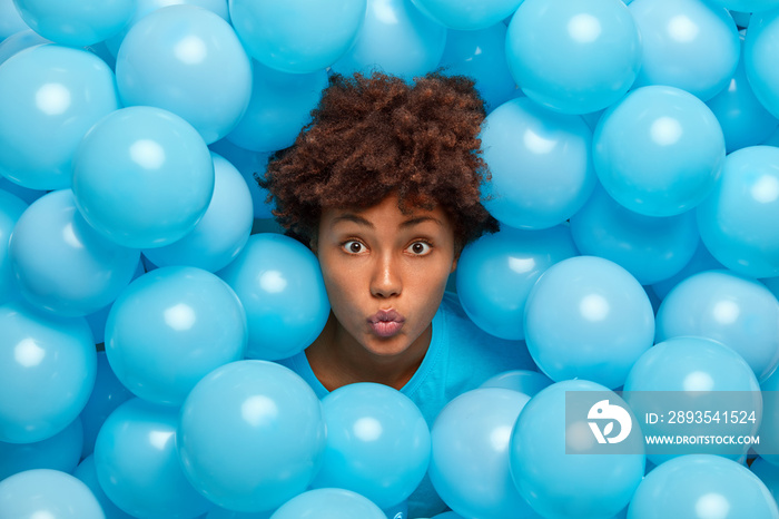 Photo of young Afro American woman keeps lips rounded poses around blue inflated balloons during fes