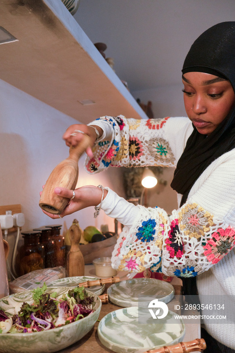 Woman wearing hijab seasoning salad in kitchen