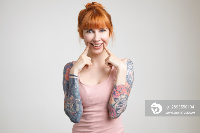 Joyful green-eyed young redhead woman with tattoos holding index fingers on her cheeks while smiling