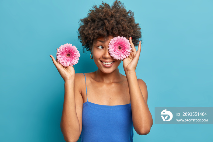 Positive dark skinned young woman covers eyes with pink gerberas smiles happily has curly bushy hair
