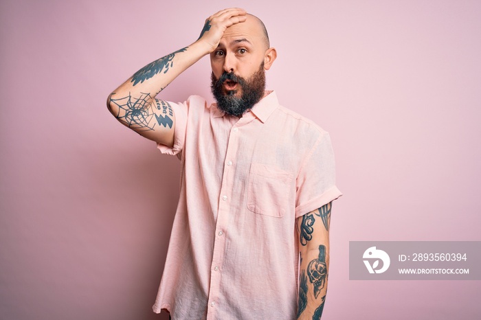 英俊的秃头男子，留着胡子，身上有纹身，穿着粉色背景的休闲衬衫，令人惊讶