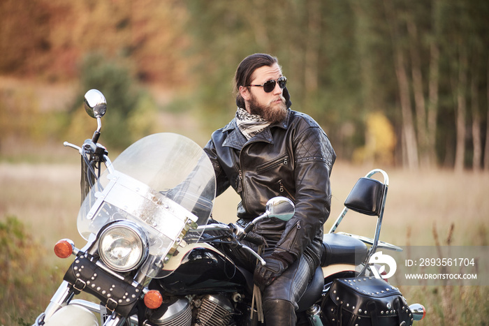 年轻的大胡子摩托车手，穿着黑色皮衣，戴着深色太阳镜，坐在黑色闪亮模式下
