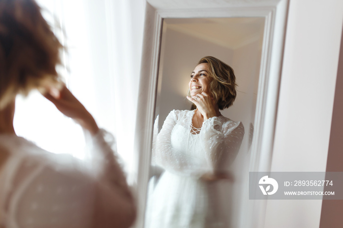 Bride in elegant white dress looking in the mirror. The bride smiles and poses in front of the mirro