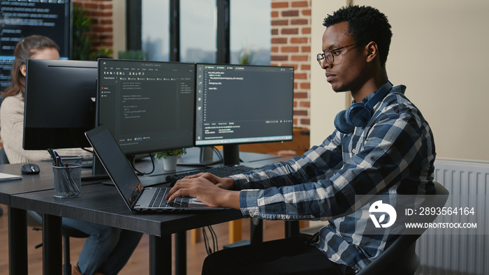 Programer thinking while touching beard and fixing glasses while typing on laptop sitting at desk wi