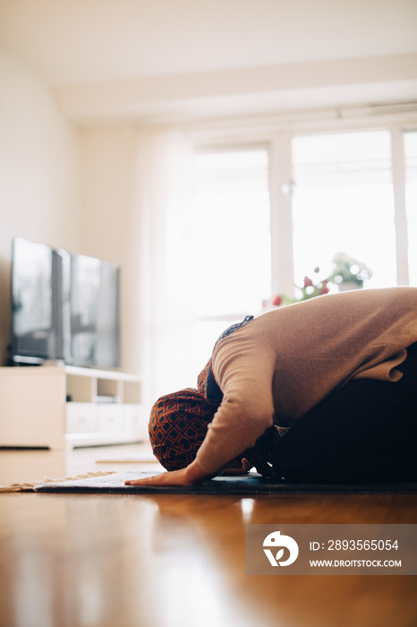 Woman wearing hijab praying on carpet in living room at home