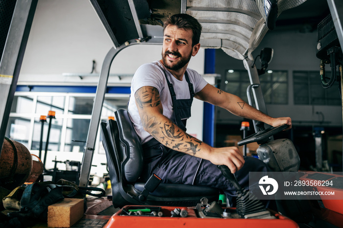 Smiling bearded tattooed hardworking employee in overalls sitting in vehicle and driving it.