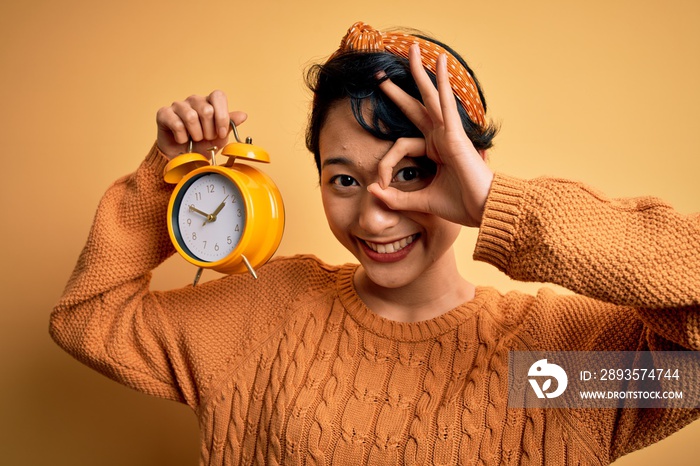 Young beautiful chinese woman holding vintage alarm clock over isolated yellow background with happy