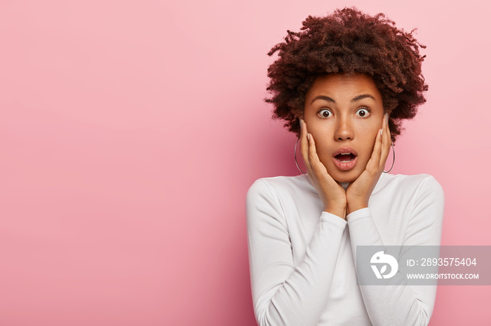 Headshot of stupefied emotional dark skinned woman gazes at camera with shock, keeps jaw dropped fro