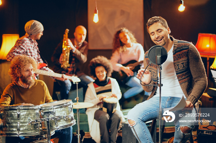 Man singing and sitting on chair while his band playing instruments in background. Home studio inter