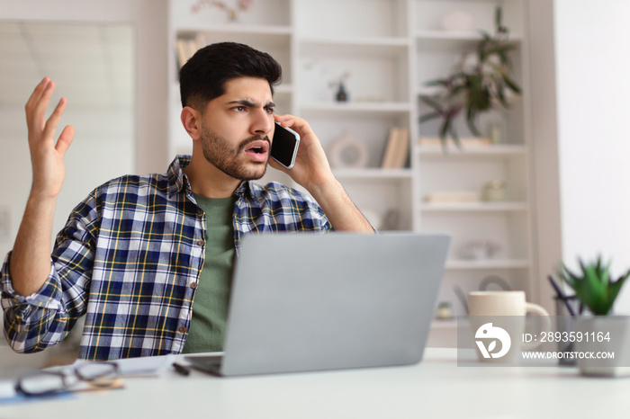 Confused Arab man talking on mobile phone using laptop