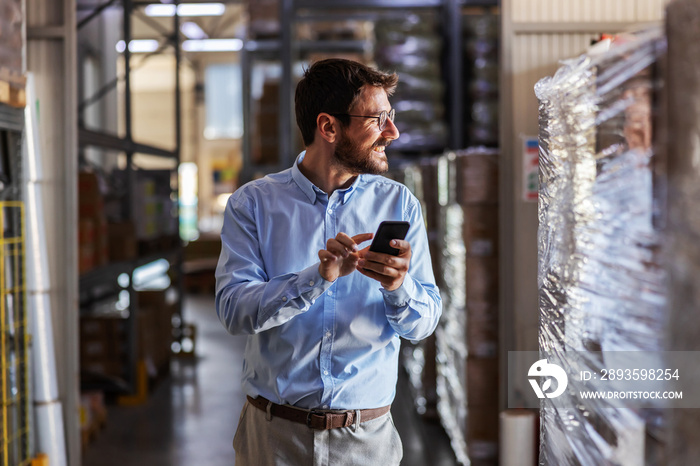 Young smiling attractive bearded businessman standing in warehouse and using smart phone.