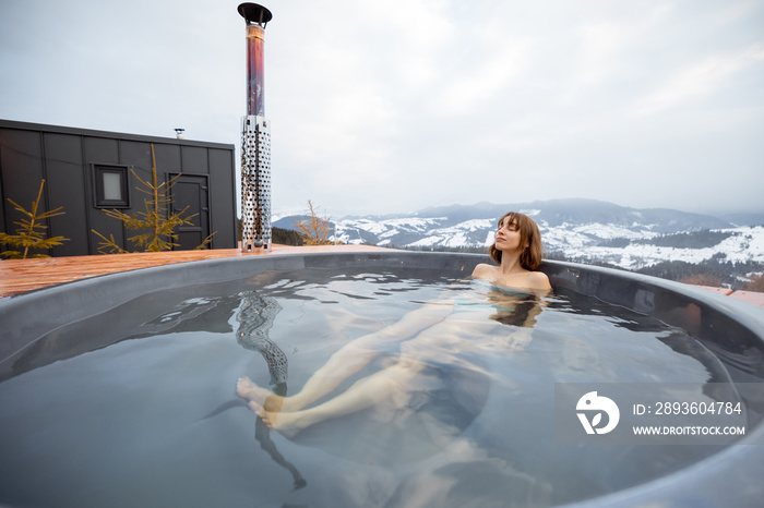 Woman swims in hot bath while resting at small modern house in the mountains on winter. Concept of r